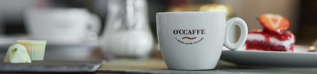 Vit O'Ccaffè-kopp på ett bord med bakverk i bakgrunden