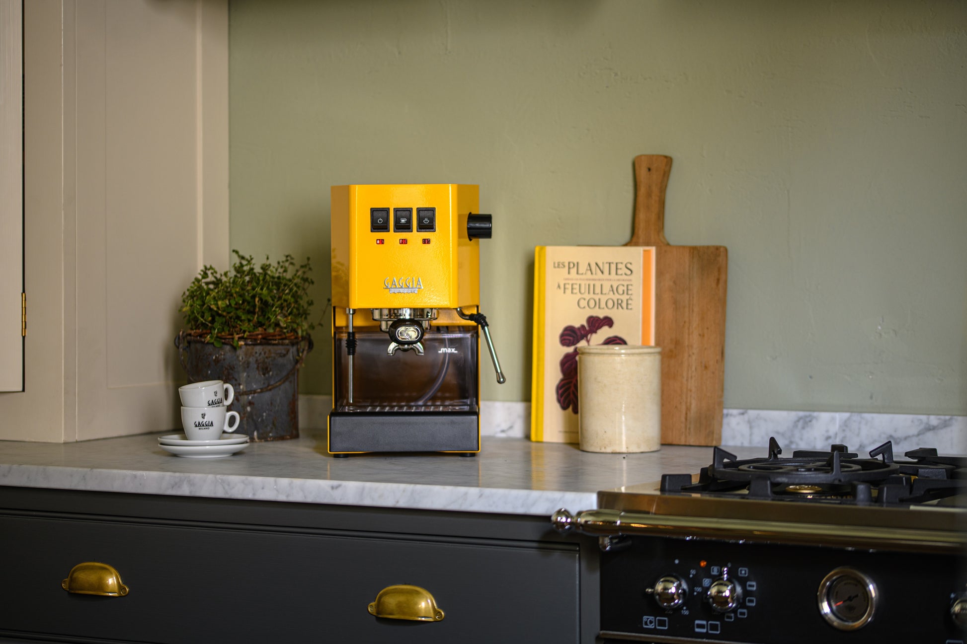 Gaggia Classic E24 espressomaskin gul på bänk med espressokoppar, serveringsbricka och en kruka med örter